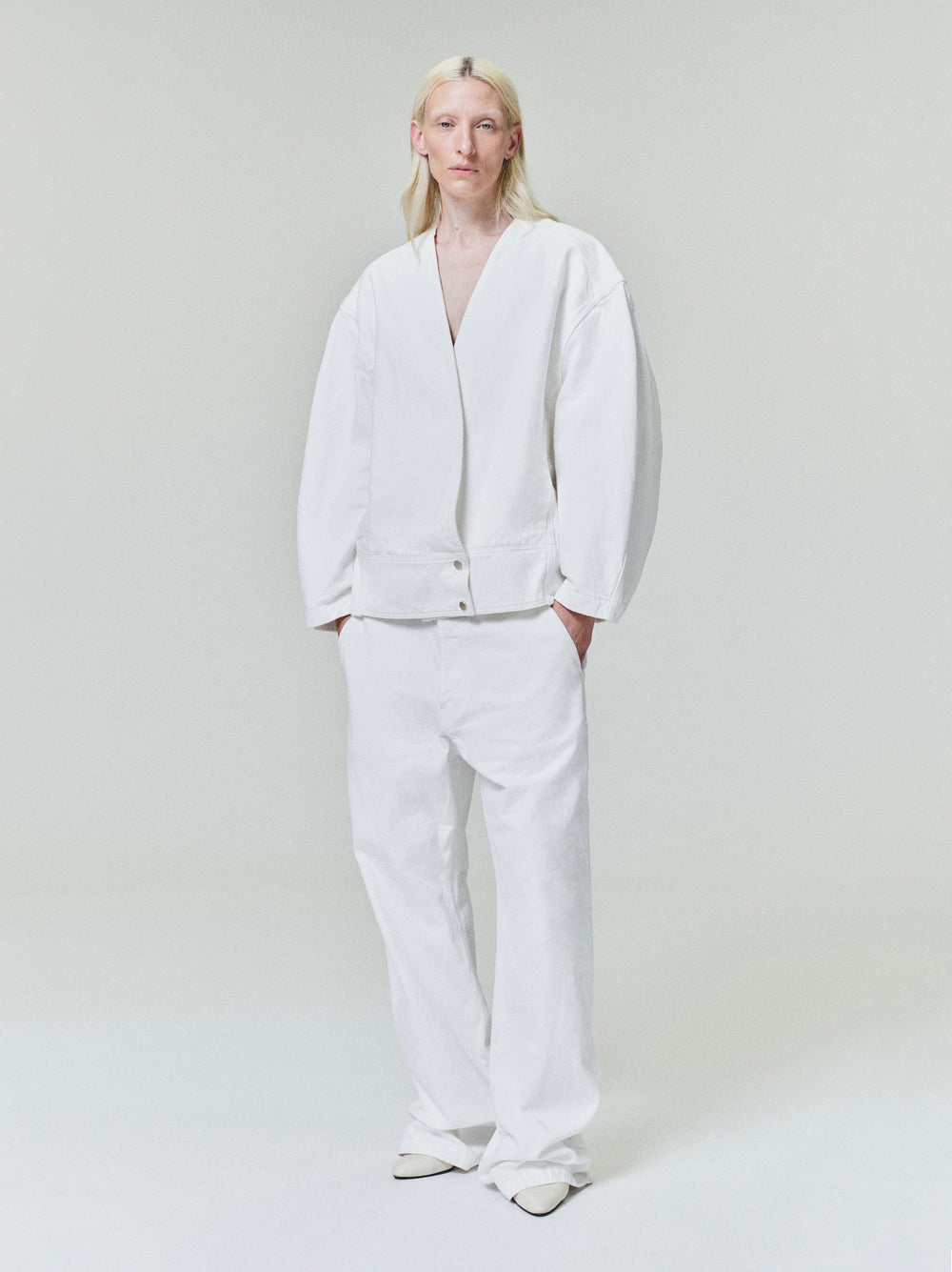 Person with long light blonde hair wearing oversized white jacket and matching loose white pants on light gray background