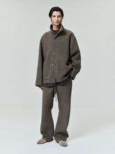 Man wearing a loose brown button-up jacket with large front pockets and matching wide-legged pants standing against a plain backdrop