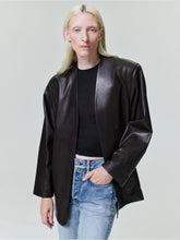 Person with straight pale blonde hair wearing oversized black leather jacket and light blue jeans on plain background