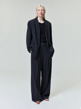 Person with pale blonde hair wearing loose-fitting black blazer and wide-leg black trousers on light gray background