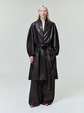 Person with long pale blonde hair wearing a belted black leather coat and wide black pants on plain background