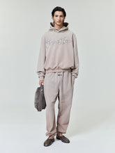 Young man wearing a beige hoodie with "Fear of God" script and matching loose sweatpants, holding a gray bag