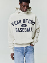 Man wearing an off-white hoodie with bold black "Fear of God Baseball" text and a front kangaroo pocket paired with light blue jeans