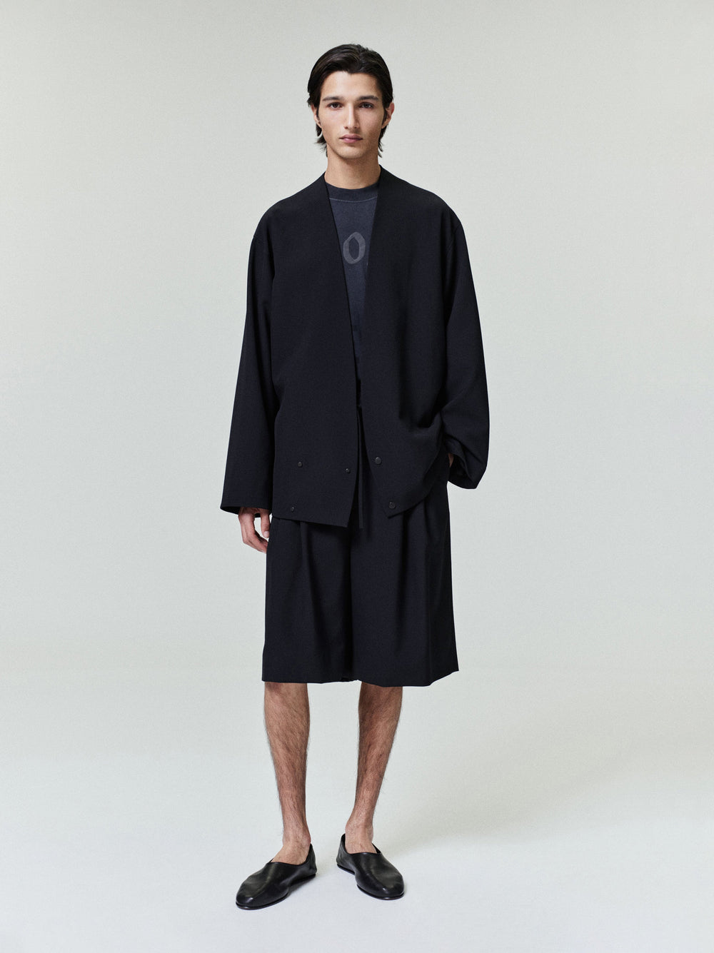 Model wearing a black Wool Ripstop Relaxed Cropped Lapelless Jacket paired with matching shorts and black slip-on shoes in a minimalist studio setting.