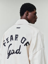 Man wearing an off-white oversized jacket with bold black "Fear of God" lettering on the back against a plain background