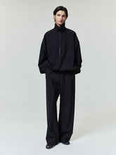 Man wearing an oversized black pullover with a high collar and wide black trousers on a plain background