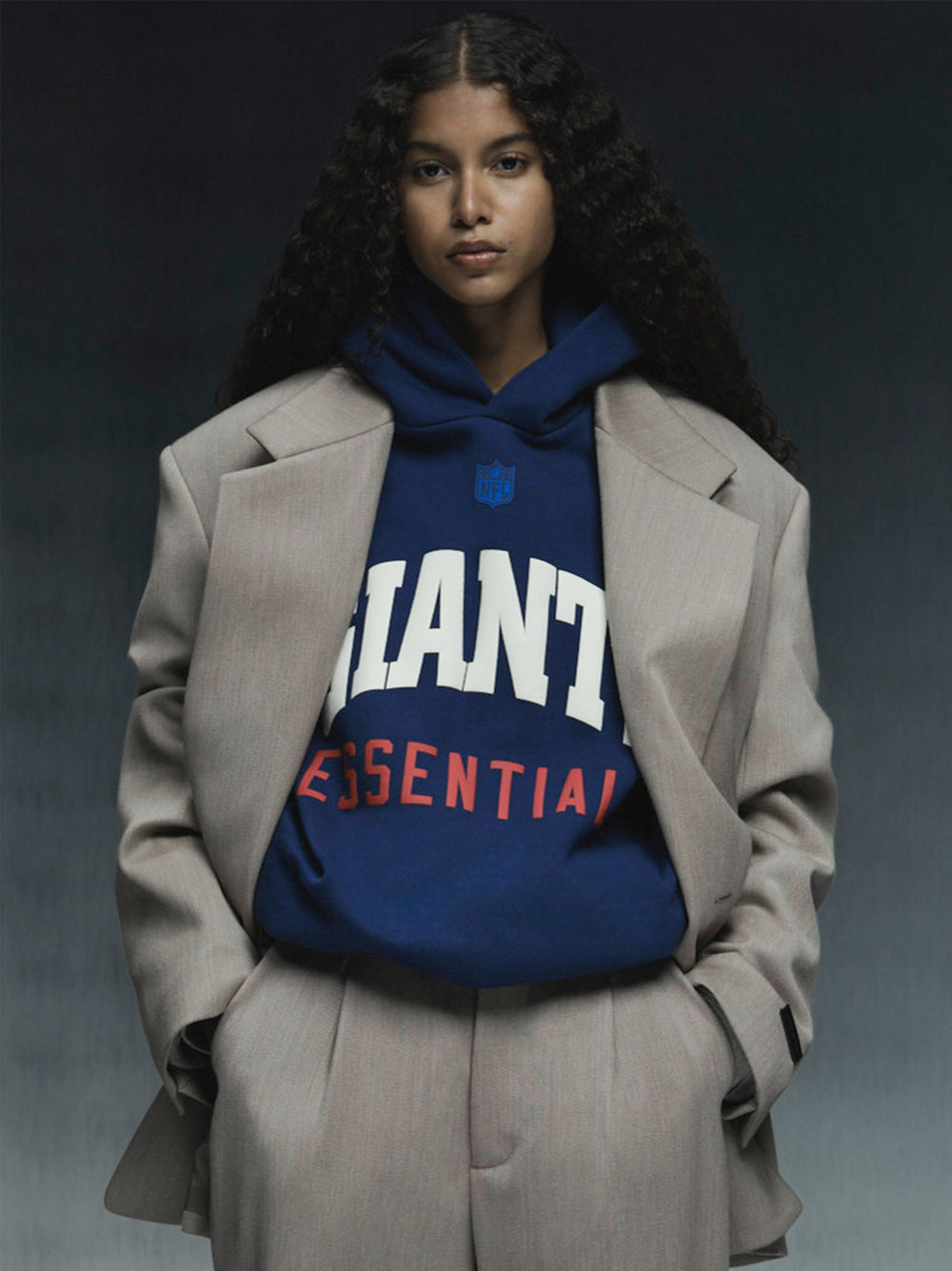 Model wearing blue Giants Sport Hoodie with bold white and red text under beige oversized suit jacket against dark background