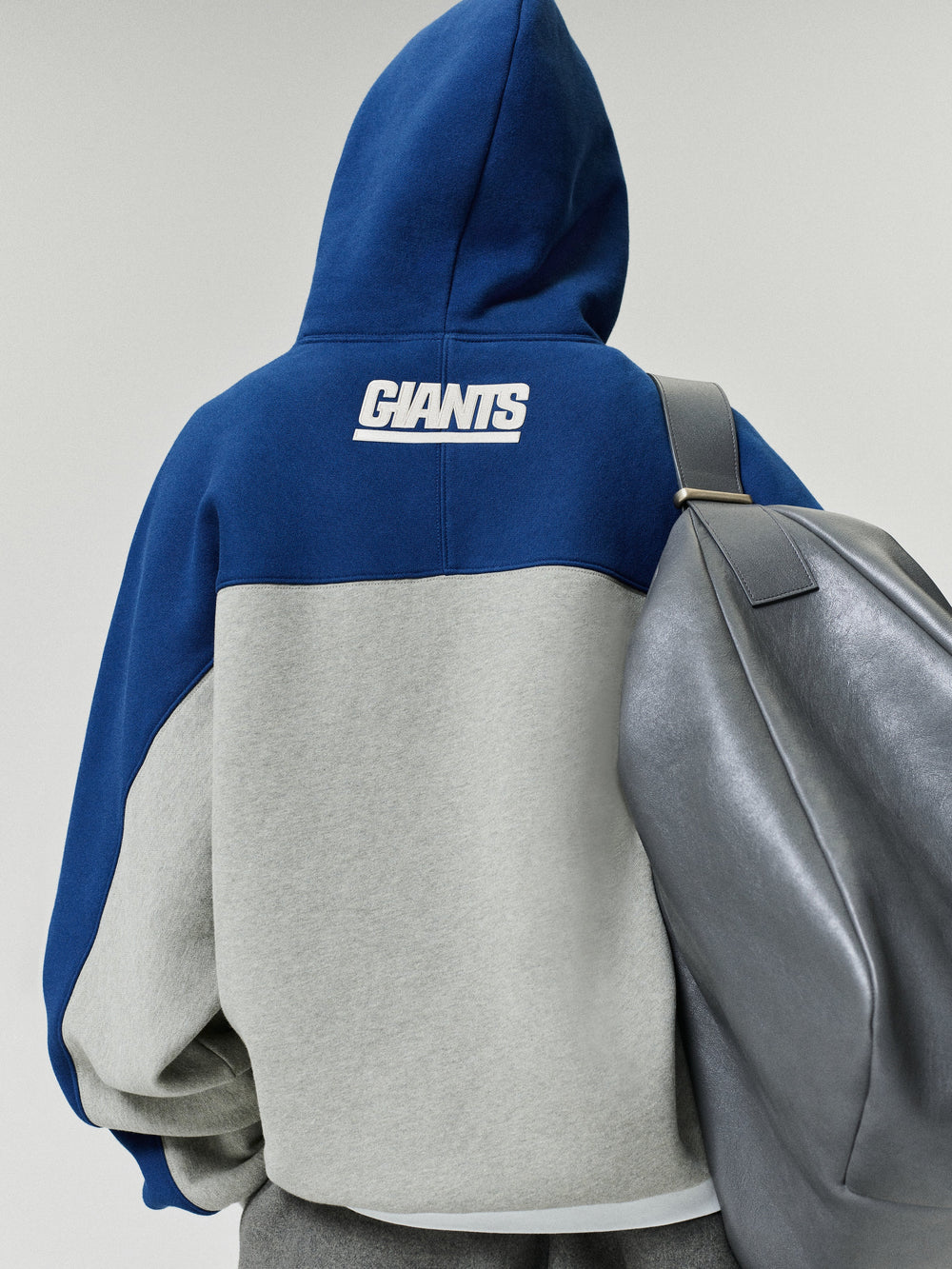 Giants 90s Relaxed Hoodie shown from the back in blue and gray with a white Giants logo and color blocked fleece body styled with a gray backpack as a Tops fashion view