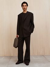 Young person with sleek dark hair wearing a loose black suit holding a gray bag against a light wood wall
