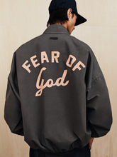 Person wearing a dark oversized jacket with "Fear of God" text printed in large letters on the back against a light wood background