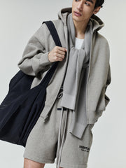 Young man wearing a light gray zip-up hoodie with matching drawstring shorts featuring small black text, a white t-shirt underneath, a textured gray scarf around the neck, and carrying a large black fabric tote bag.