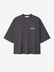 Merch Tee in dark iron grey featuring a relaxed oversized fit with dropped shoulders and small cream text "ALL VALLEY ESSENTIALS" on the chest for a minimalist casual top.