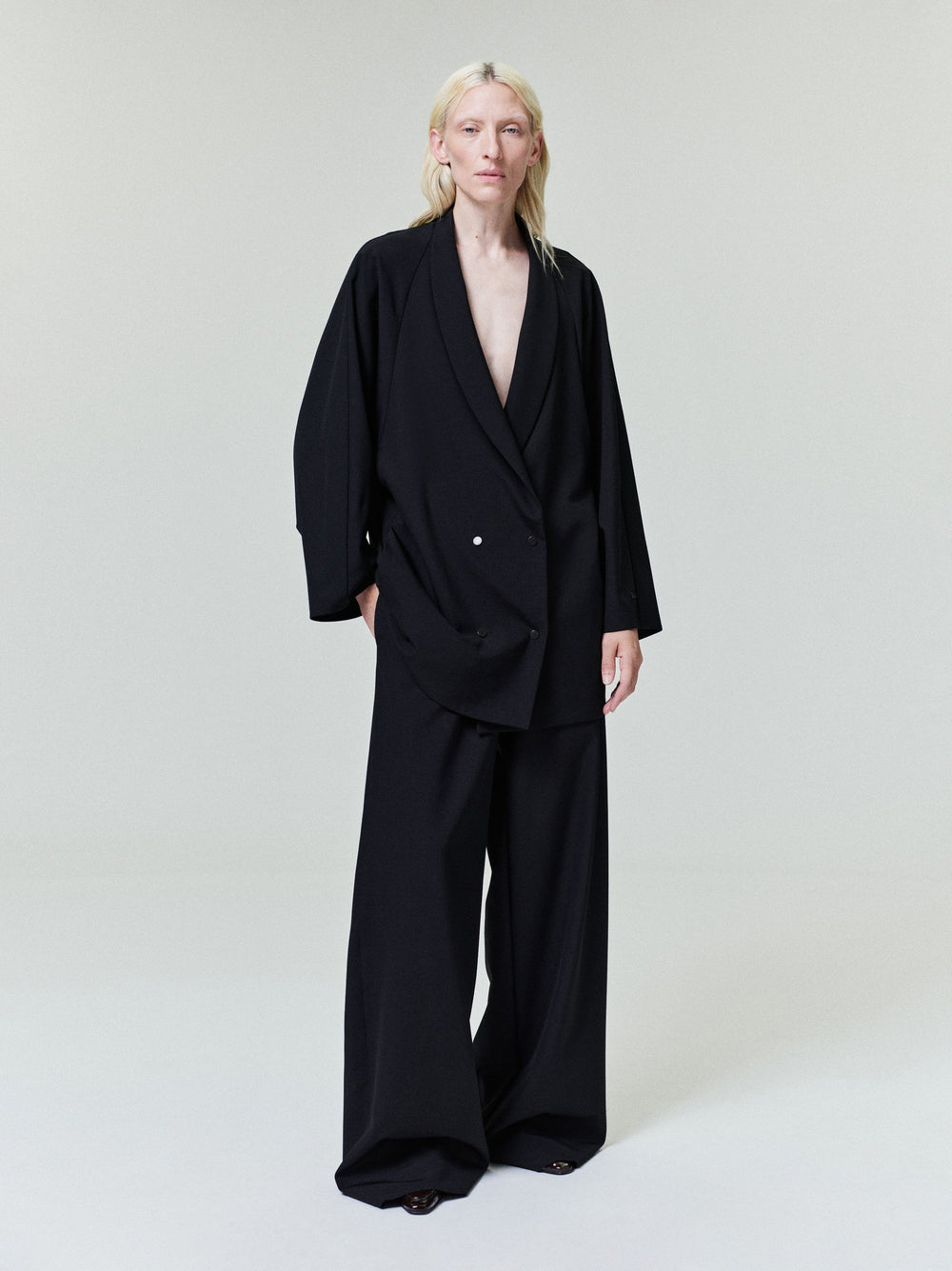 Person with pale blonde hair wearing oversized black double-breasted blazer and wide-leg black trousers on light background