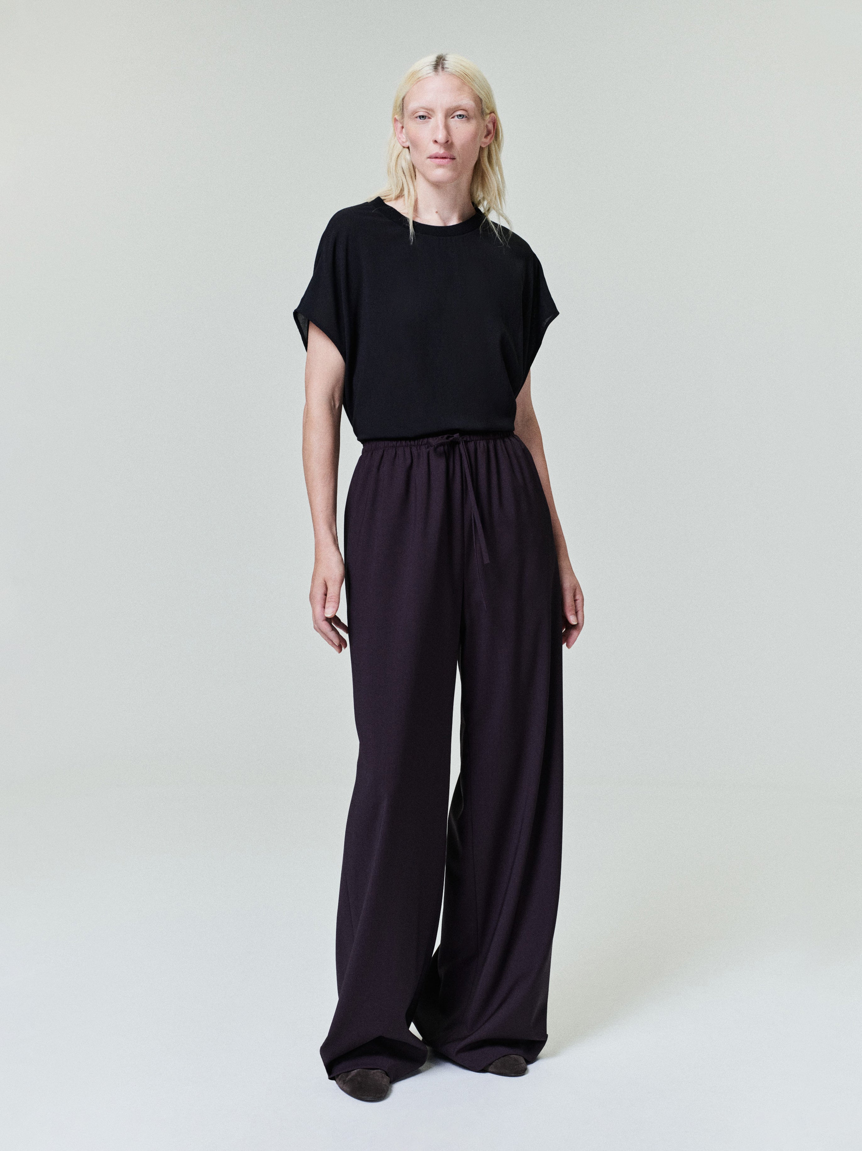 Person with pale blonde hair wearing loose black short-sleeve top and wide-leg dark purple pants on plain light background