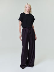 Person with pale blonde hair wearing loose black short-sleeve top and wide-leg dark purple pants on plain light background