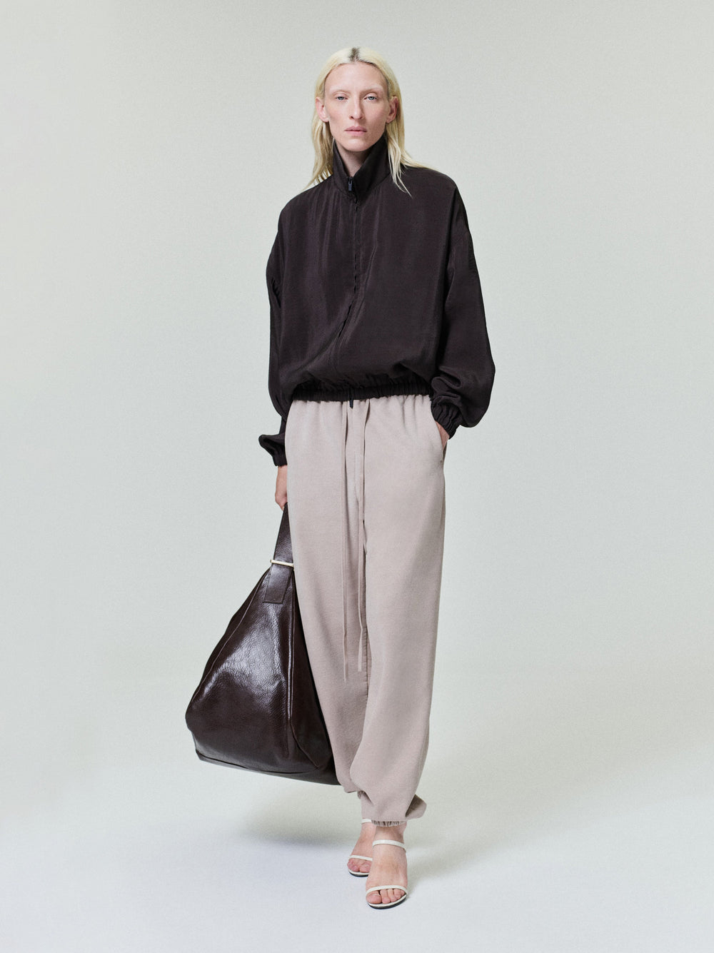 Person with pale blonde hair wearing loose black zip-up jacket, beige drawstring pants, and white heels carrying large dark bag
