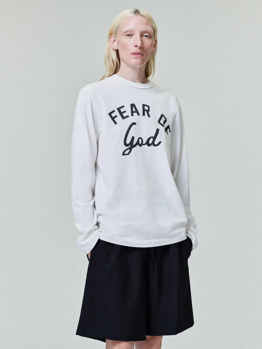 Person with pale blonde hair wearing white long-sleeve shirt with "Fear of God" text and black shorts on light background
