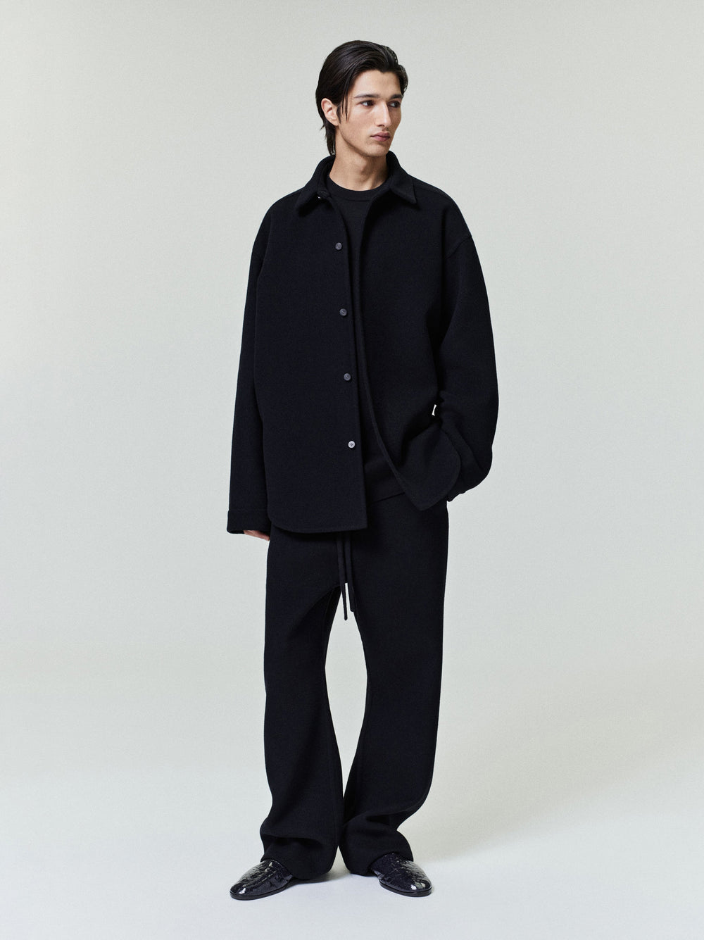 Young man wearing a relaxed black button-up shirt jacket with long sleeves and matching loose black trousers standing on a light background