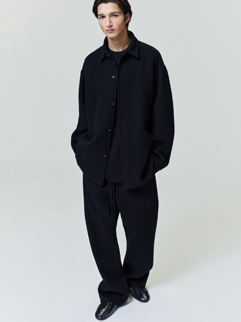 Man wearing an oversized black button-up shirt jacket with relaxed sleeves and wide black trousers against a plain backdrop