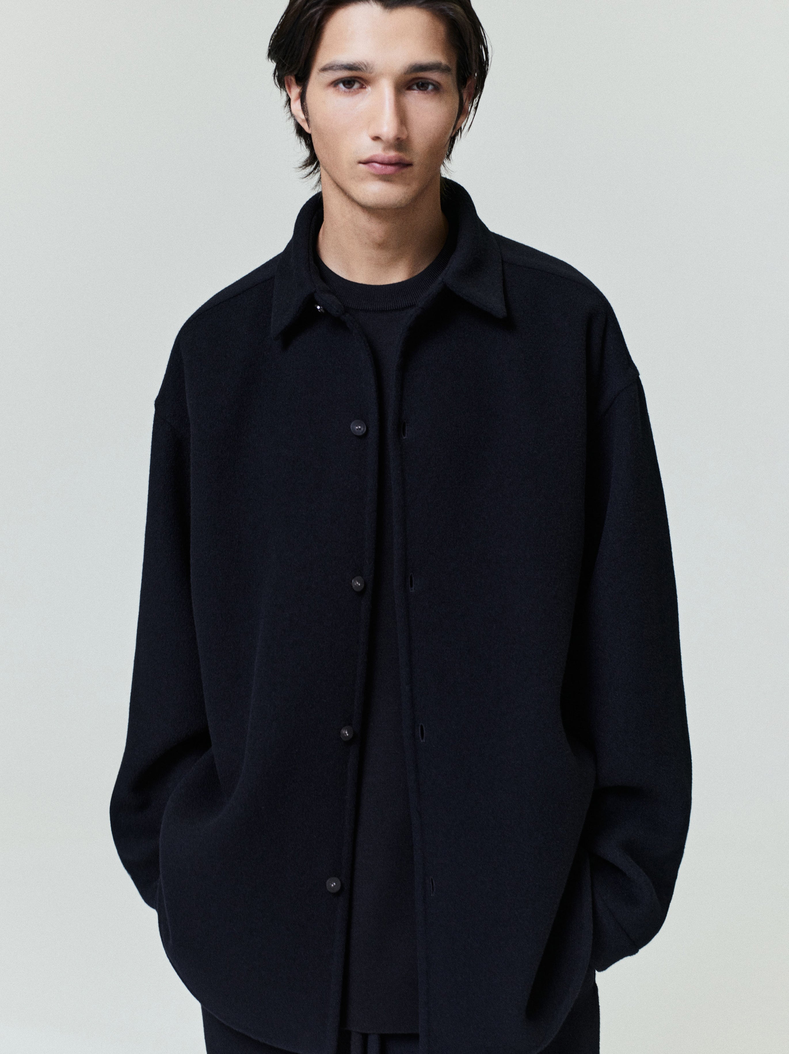 Young man wearing a relaxed black button-up shirt jacket with dropped shoulders and side pockets over a black shirt