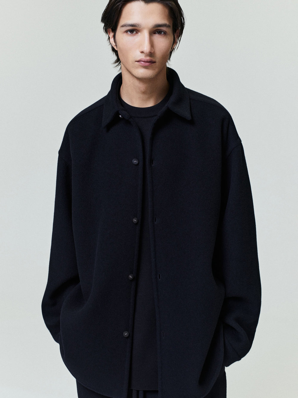 Young man wearing a relaxed black button-up shirt jacket with dropped shoulders and side pockets over a black shirt