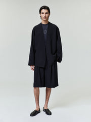 Model wearing a black Wool Ripstop Relaxed Cropped Lapelless Jacket paired with matching shorts and black slip-on shoes in a minimalist studio setting.