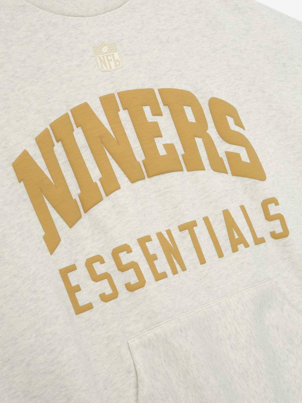 Close-up of the front of a Forty Niners Sport Crewneck in light gray, featuring large gold "NINERS" text arched above "ESSENTIALS" lettering and a small NFL shield logo, with visible fabric texture and a kangaroo pocket.