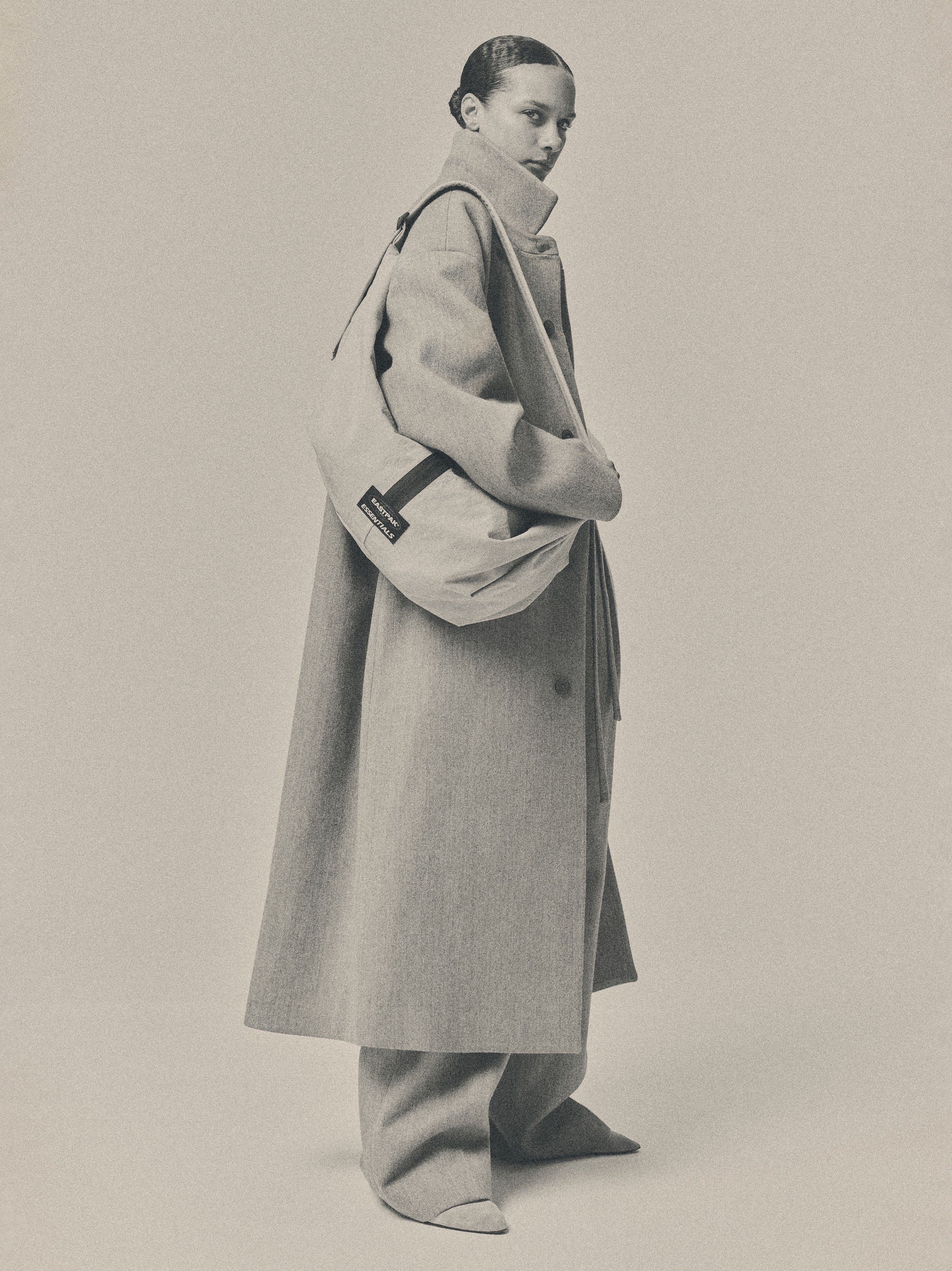 Essentials Shell Bag worn over the shoulder by a model in a long coat and relaxed trousers against a softly lit monochrome studio backdrop