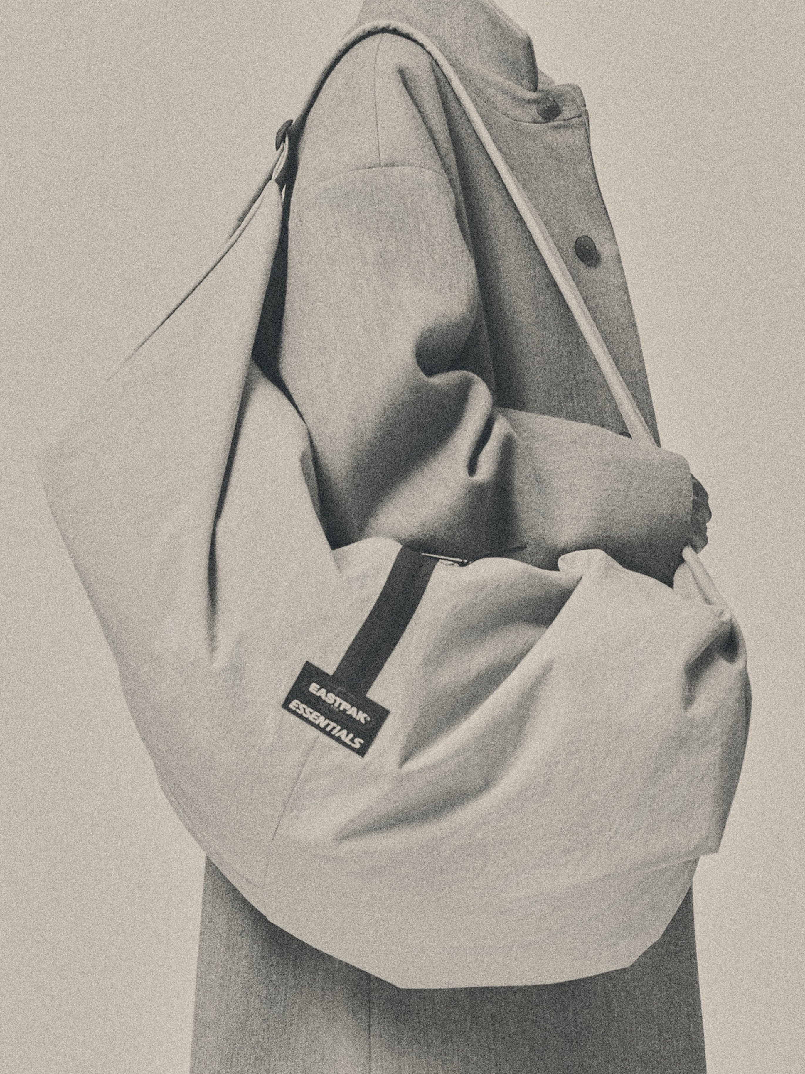 ESSENTIALS SHELL BAG LARGE worn over the shoulder in light beige with a spacious curved design black woven label and a model in a neutral coat