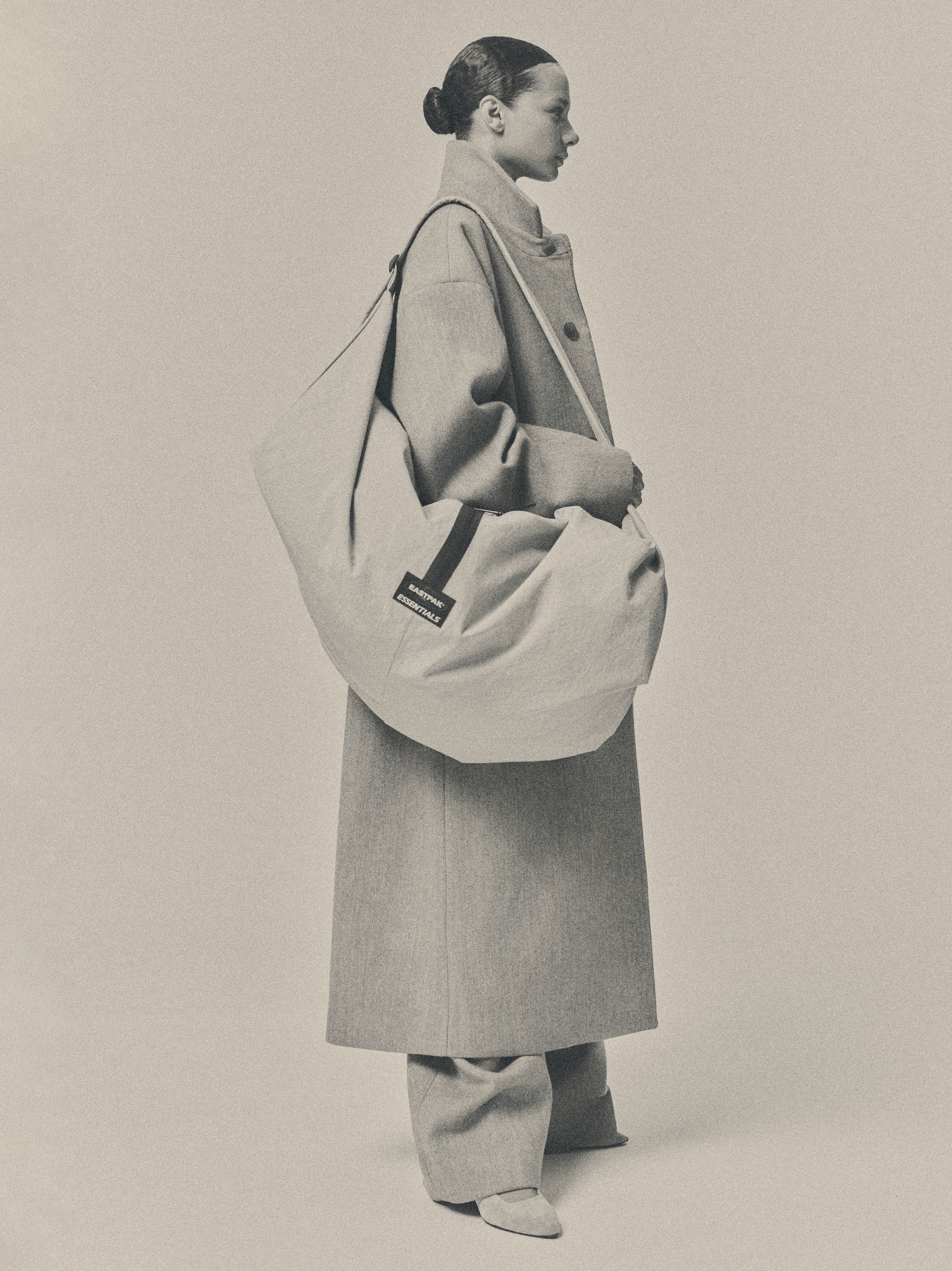 ESSENTIALS SHELL BAG LARGE in light beige carried over the shoulder by a model wearing a long neutral coat with its curved silhouette and woven logo label visible