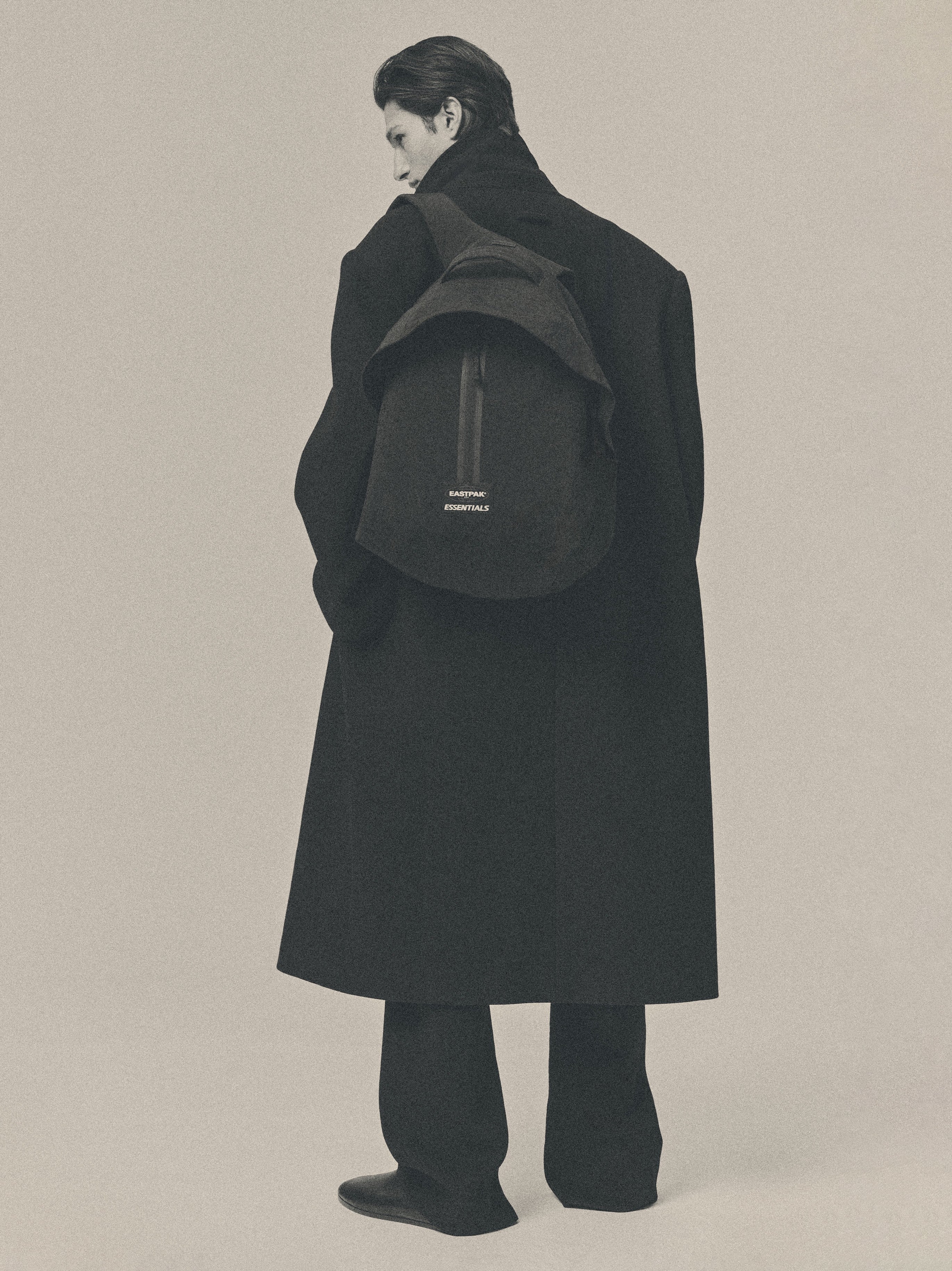 Essentials Backpack in black worn by a model over a long dark coat showing its rounded silhouette and small logo against a muted neutral studio background