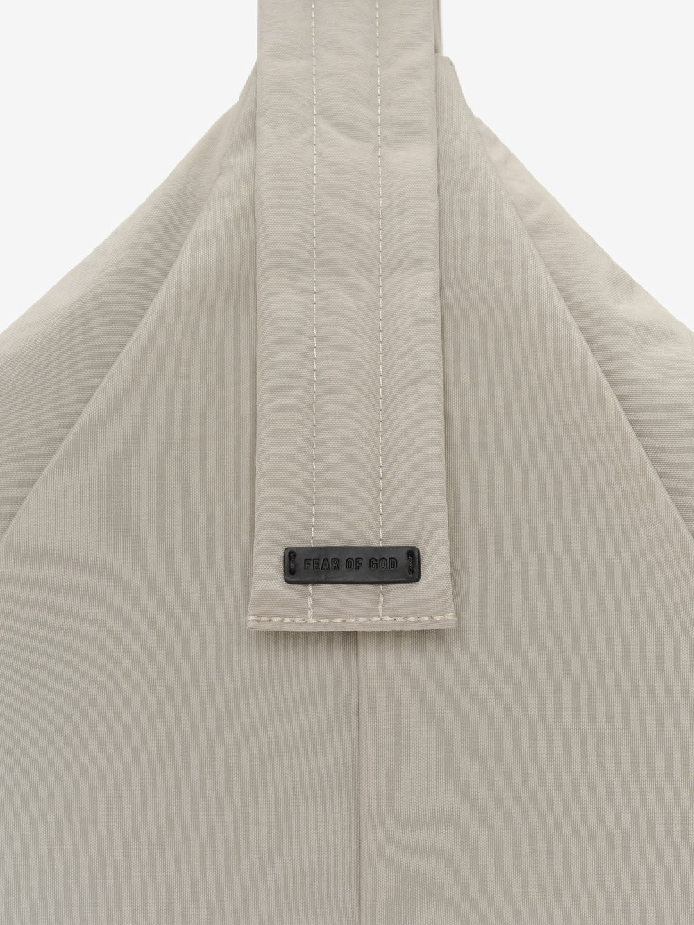 ESSENTIALS SHELL BAG LARGE close up in light beige showing a stitched central strap and Fear of God logo label on smooth fabric against a white background