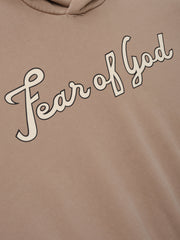 Close-up of a dune-colored hoodie with cream "Fear of God" cursive script printed diagonally across the chest