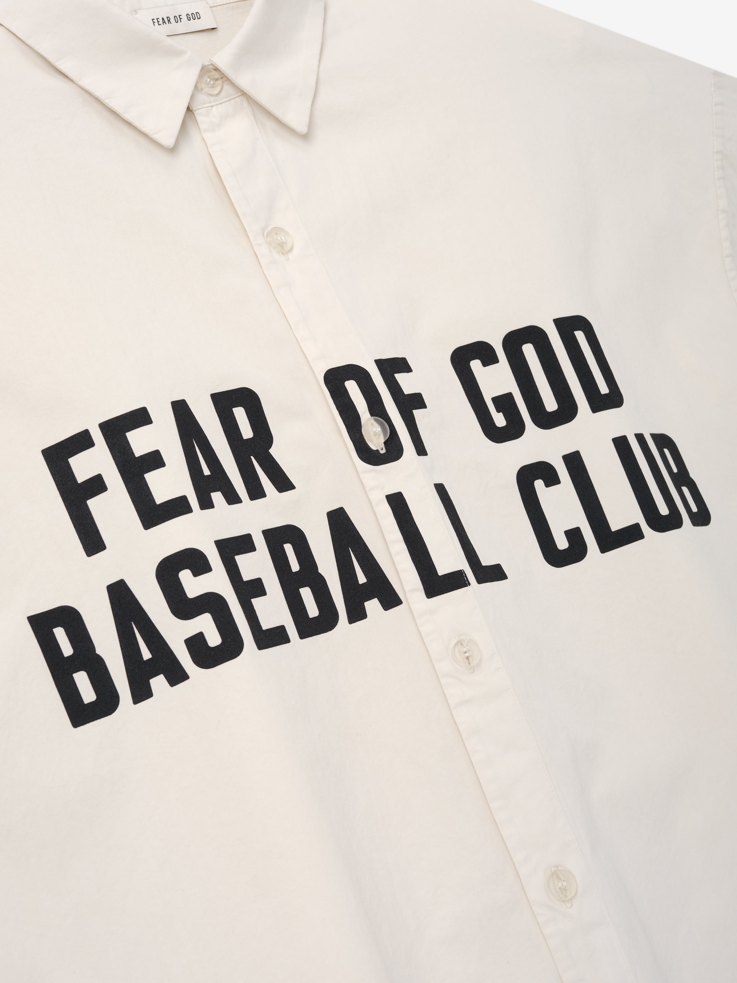 Close-up of a natural-colored shirt front with bold black "Fear of God Baseball Club" text and button-up detail
