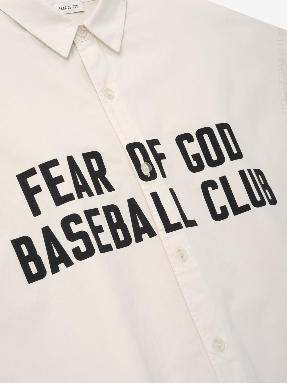 Close-up of a natural-colored shirt front with bold black "Fear of God Baseball Club" text and button-up detail