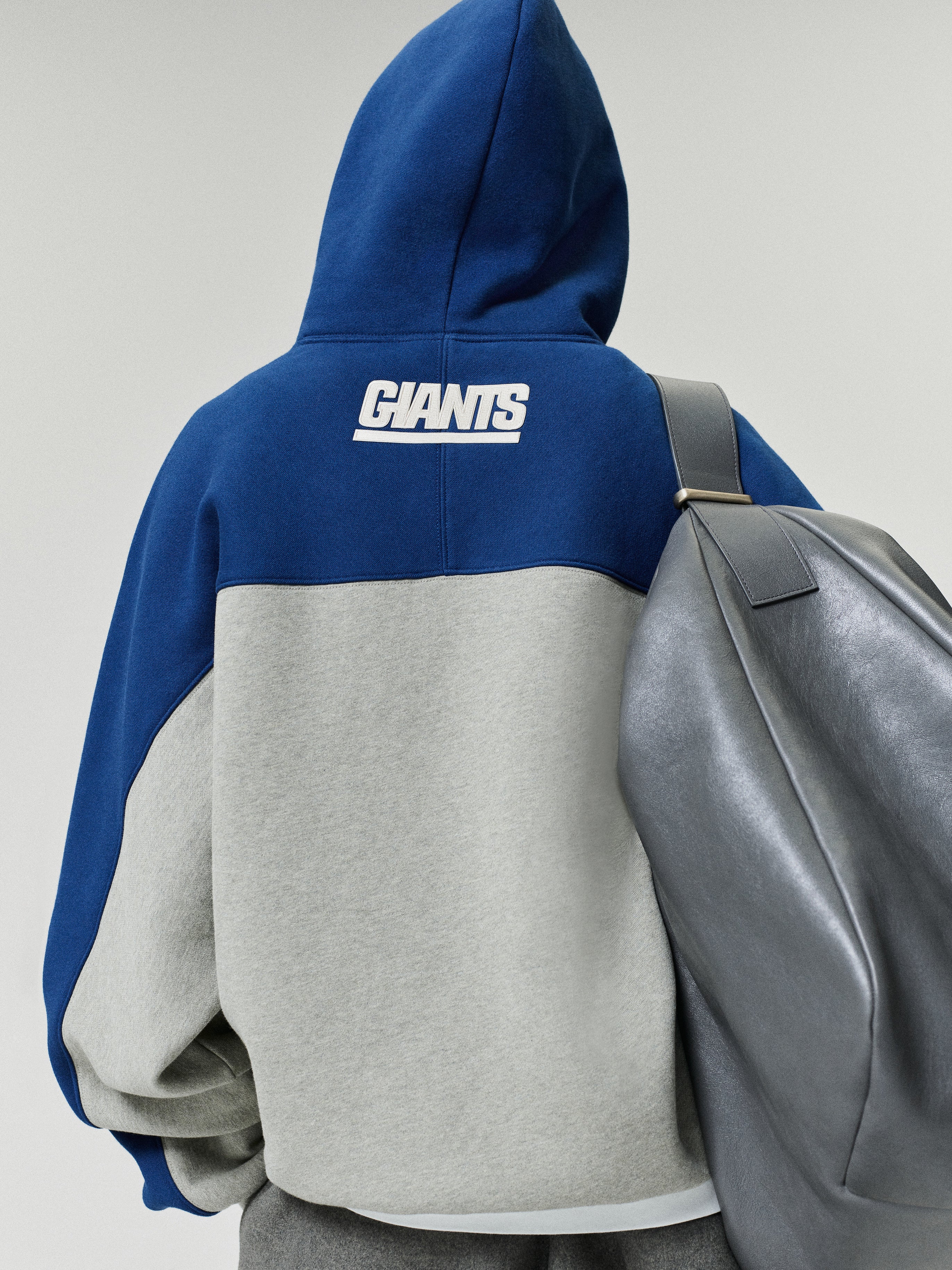 Giants 90s Relaxed Hoodie shown from the back in blue and gray with a white Giants logo and color blocked fleece body styled with a gray backpack as a Tops fashion view