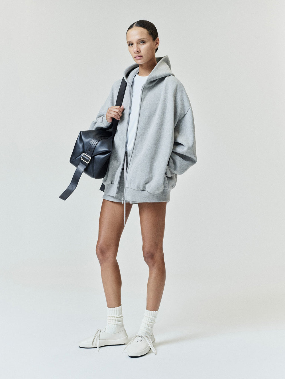 Womens Embossed FW26 Cropped Zip-Up Hoodie in light gray styled with a black shoulder bag and white sneakers Fear of God Tops
