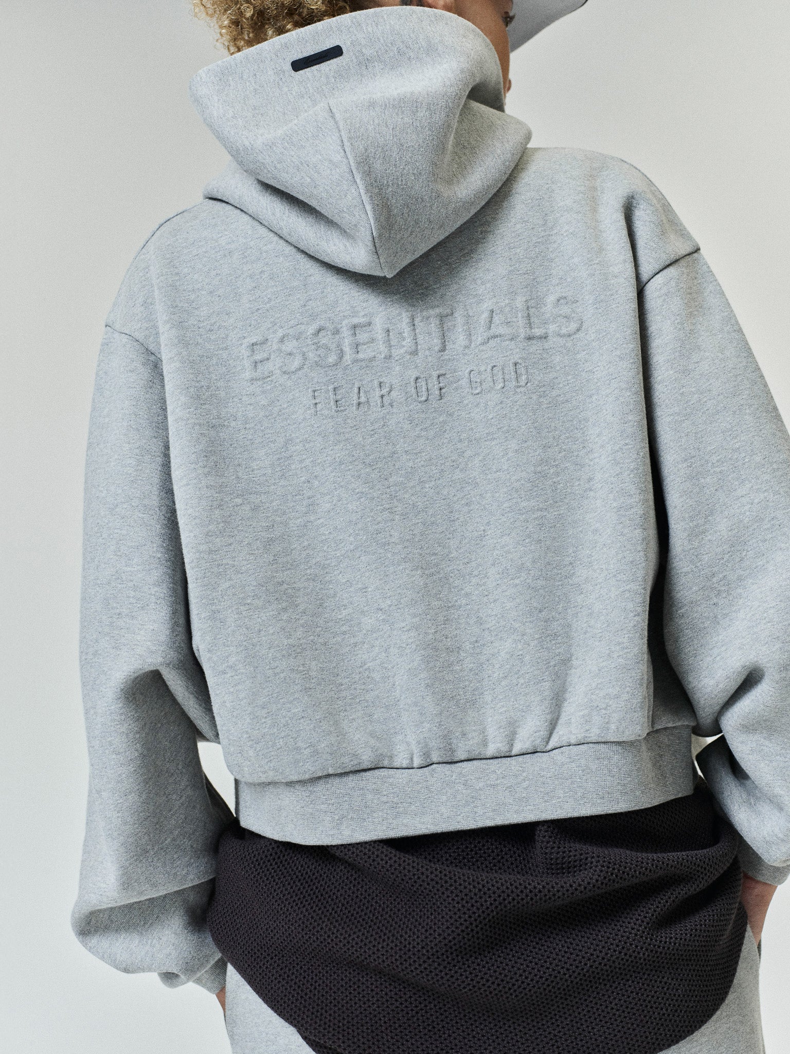 Womens Embossed FW26 Cropped Zip-Up Hoodie in light gray shown from the back with embossed ESSENTIALS Fear of God lettering hood and cropped hem Tops