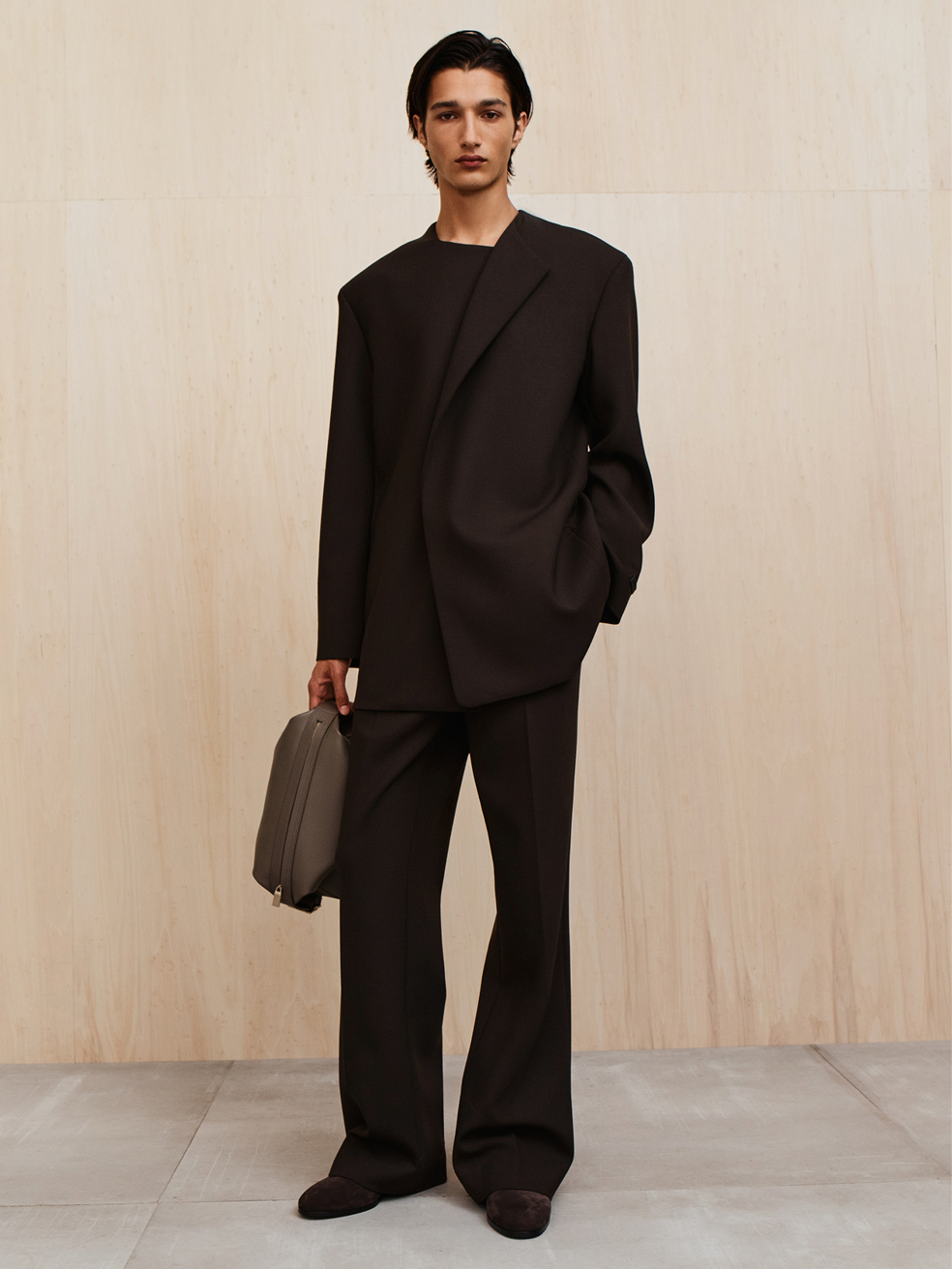 Young person with sleek dark hair wearing a loose black suit holding a gray bag against a light wood wall