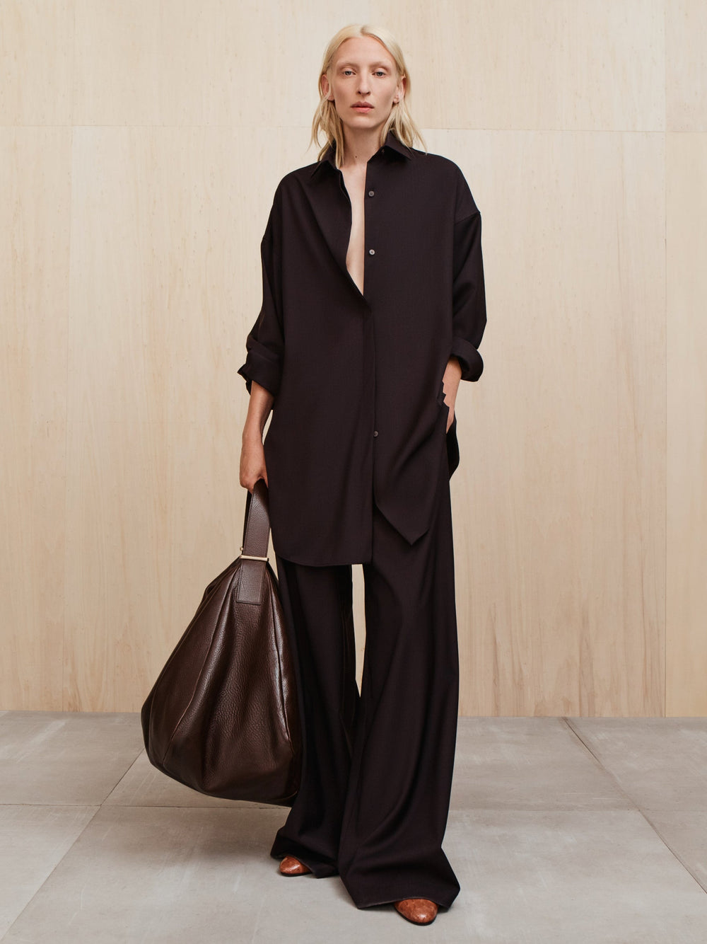 Person with light blonde hair wearing a loose black button-up shirt and wide black pants holding a large dark brown bag