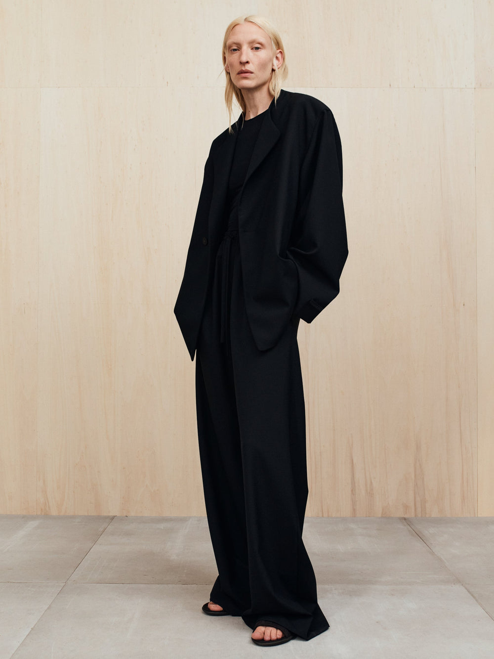 Person with pale blonde hair wearing an oversized black suit with wide-leg pants standing against a light wood wall