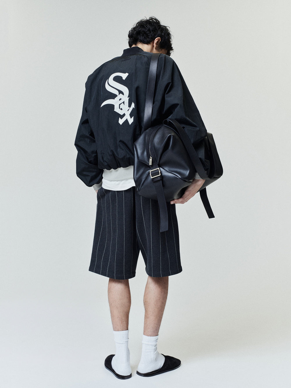 Model wearing White Sox Half Zip Pullover in black with a large white logo on the back, paired with pinstriped shorts, white socks, black slides, and carrying a black leather backpack.
