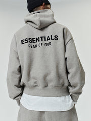 Person wearing a light gray oversized hoodie with black "ESSENTIALS FEAR OF GOD" text on the back, paired with matching pants and a black knit beanie.