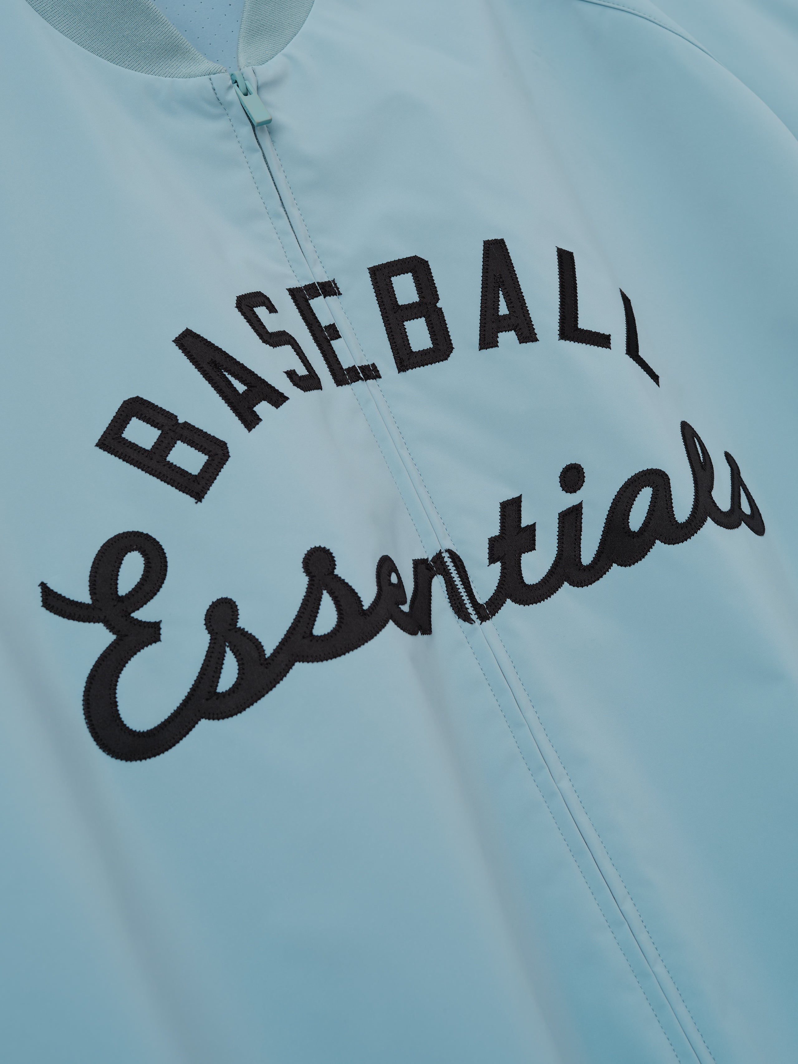 MLB Full Zip Bomber in muted blue featuring a centered black embroidered "Baseball Essentials" script across the chest, designed as a lightweight outerwear top.