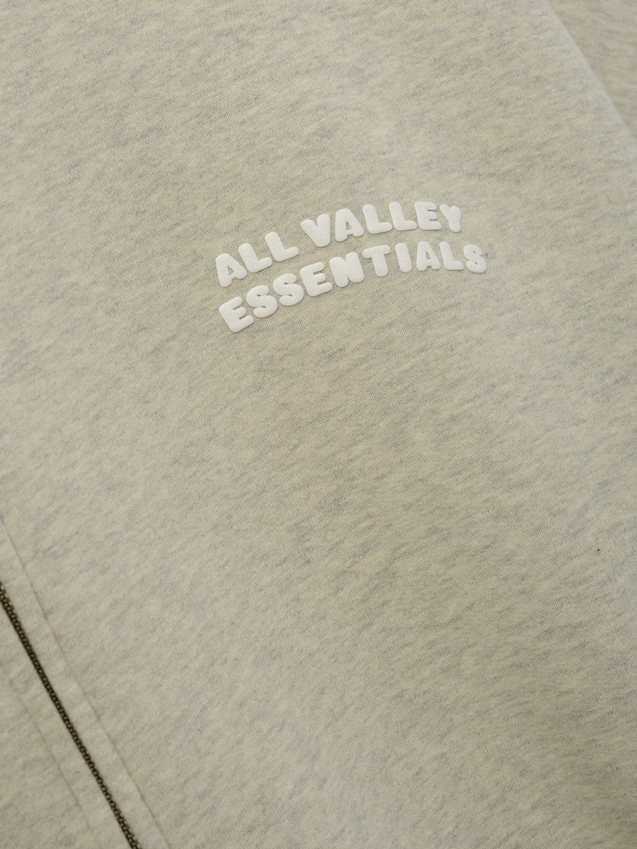 90'S Zip-Up Hoodie in a soft washed vanilla heather fabric featuring close-up of raised white "ALL VALLEY ESSENTIALS" lettering on the chest near the zipper seam.