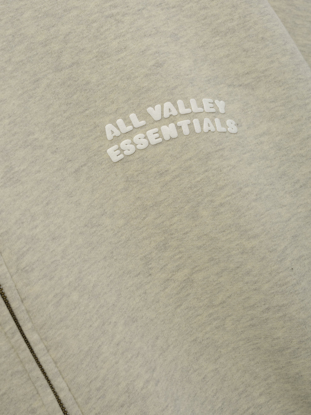 90'S Zip-Up Hoodie in a soft washed vanilla heather fabric featuring close-up of raised white "ALL VALLEY ESSENTIALS" lettering on the chest near the zipper seam.