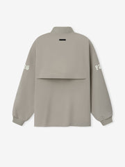 Half Zip Track Jacket in a soft muted taupe featuring long sleeves with white printed lettering on each arm, a high collar, and a unique layered back panel for modern outerwear style.