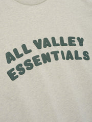 Merch Tee in light heathered neutral shade featuring soft fabric with bold dark green "ALL VALLEY ESSENTIALS" text curved across the front for a relaxed casual top.
